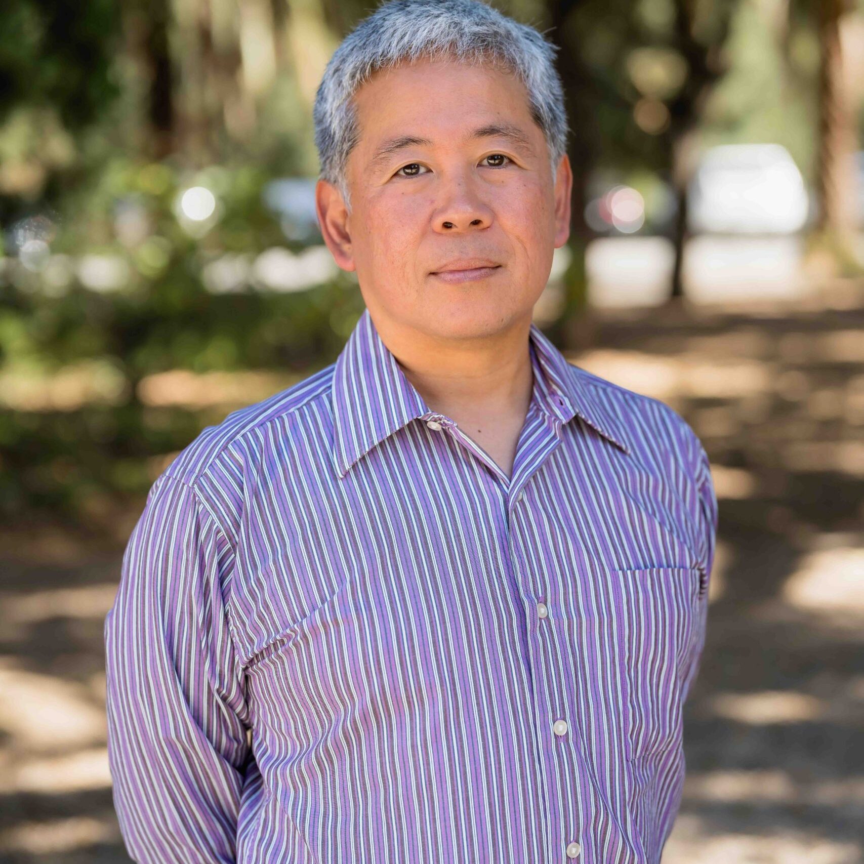 Hideo Mabuchi, wearing a blue and white striped button up, stands in a wooded grove, hands clasped behind his back