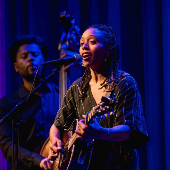 A person playing the guitar stands in a spotlight against a blue background.