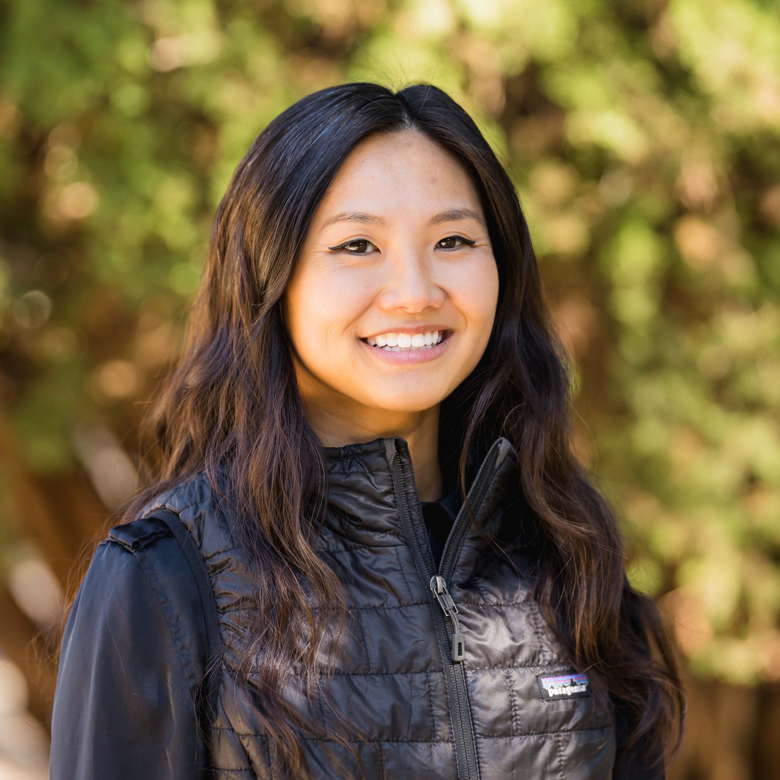 Portrait of Edi Dai who is wearing a black vest and a long sleeve shirt. They are standing in front of greenery.
