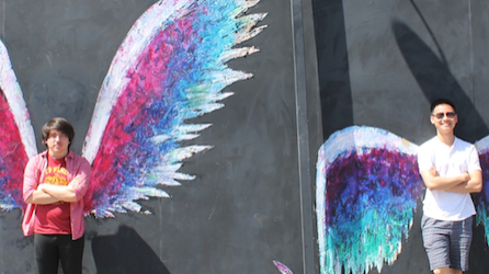 Two student pose in front of murals of large feather wings in bright colors